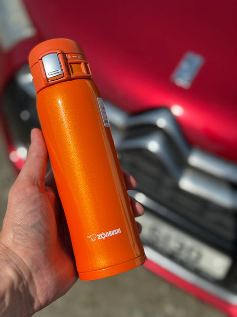 Orange thermos Zojirushi Mug SM-SHE from 480 ml held by a hand with a red vehicle in the background