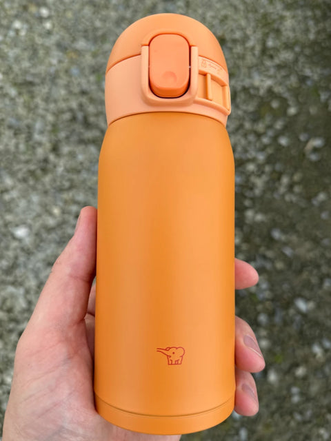 Zojirushi SM-WA orange stainless steel travel mug with flip lid, held in hand outdoors.