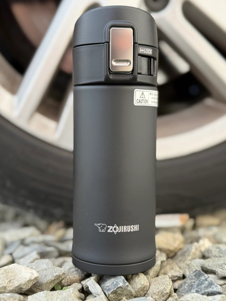 Zojirushi SM-KHE black travel mug standing on stones outdoors with car wheel in background