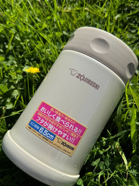 Zojirushi Food Jar SW-EAE in creamy white, 500 ml (16.9 oz), photographed outdoors on green grass with a yellow flower nearby.