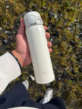 Hand holding a white Zojirushi travel mug with a one-touch locking lid, outdoors over grass and stones.