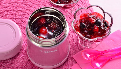 Zojirushi pink food jar filled with berry dessert, next to a glass bowl of berries and pink spoons